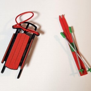 May include: A miniature red and black sled with a red rope and a pair of red skis with green and silver poles. The sled has a red seat and black frame. The skis are red with green tips.