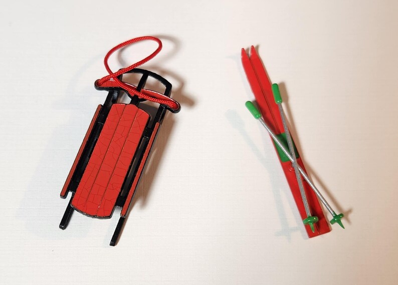 May include: A miniature red and black sled with a red rope and a pair of red skis with green and silver poles. The sled has a red seat and black frame. The skis are red with green tips.