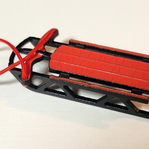 May include: A small red and black sled with a red rope. The sled has a red seat and black runners and frame. The rope is looped and attached to the front. A winter-themed decorative item.