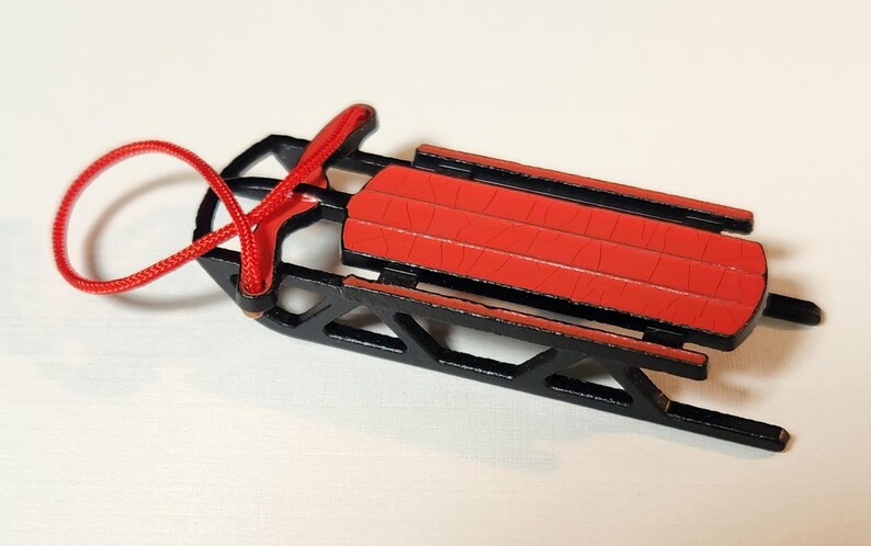 May include: A small red and black sled with a red rope. The sled has a red seat and black runners and frame. The rope is looped and attached to the front. A winter-themed decorative item.