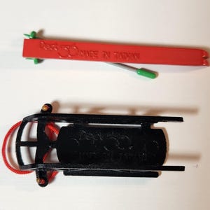 May include: Two miniature sleds are shown, one red and one black. The red sled has the text "Dept 56 MADE IN TAIWAN". The black sled has a red rope attached. These are likely Christmas decorations.