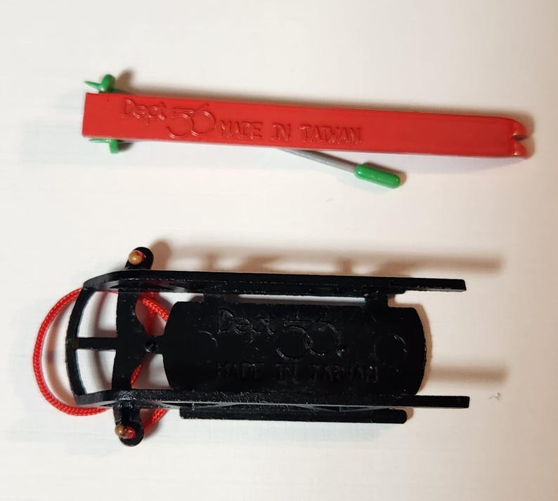 May include: Two miniature sleds are shown, one red and one black. The red sled has the text "Dept 56 MADE IN TAIWAN". The black sled has a red rope attached. These are likely Christmas decorations.