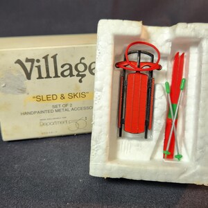 May include: A miniature red sled and skis set, presented in a white foam insert and a cream-coloured box. The box reads "Village" and "Sled & Skis". The sled and skis are hand-painted metal accessories.