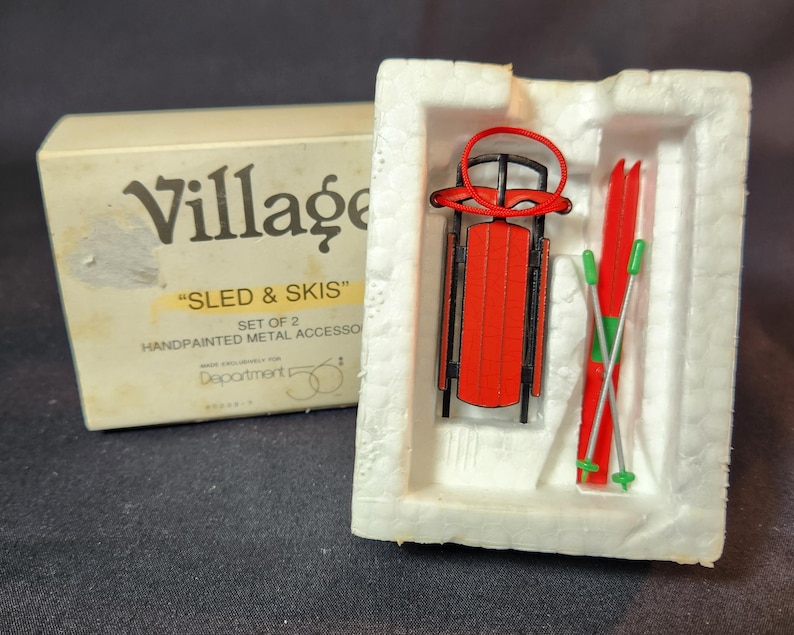 May include: A miniature red sled and skis set, presented in a white foam insert and a cream-coloured box. The box reads "Village" and "Sled & Skis". The sled and skis are hand-painted metal accessories.