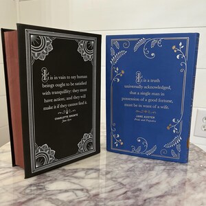 May include: Two hardcover books with decorative covers. The book on the left has a black cover with silver floral designs and the text "It is in vain to say human beings ought to be satisfied with tranquillity: they must have action; and they will make it if they cannot find it."  The book on the right has a blue cover with silver floral designs and the text "It is a truth universally acknowledged, that a single man in possession of a good fortune, must be in want of a wife."