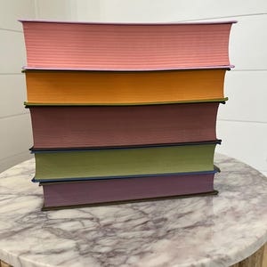 May include: A stack of six hardcover books with different colored covers. The covers are pink, orange, maroon, green, purple, and brown. The books are stacked on a round marble table with a wooden base.