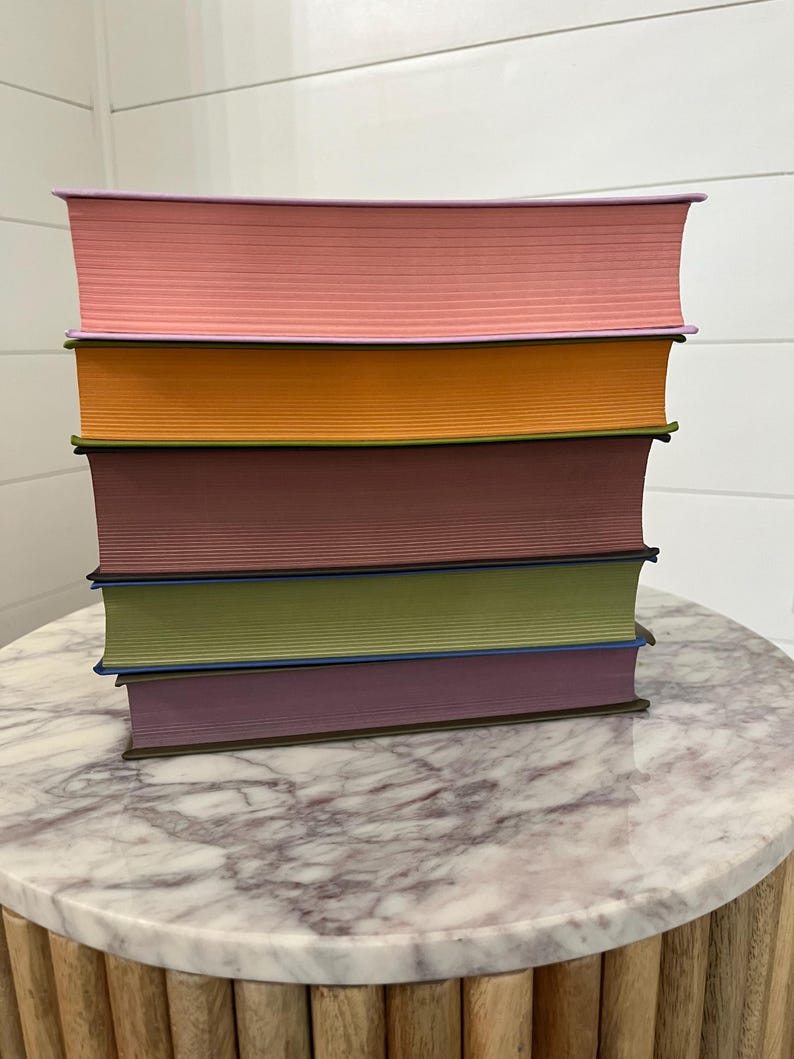 May include: A stack of six hardcover books with different colored covers. The covers are pink, orange, maroon, green, purple, and brown. The books are stacked on a round marble table with a wooden base.