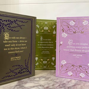 May include: Three hardcover books with gold lettering and decorative floral designs on the covers. The book on the left has a dark green cover with a quote from Emily Bront&euml;'s "Wuthering Heights." The middle book has a green cover with a quote from Jane Austen's "Sense and Sensibility." The book on the right has a light purple cover with a quote from Jane Austen's "Emma."