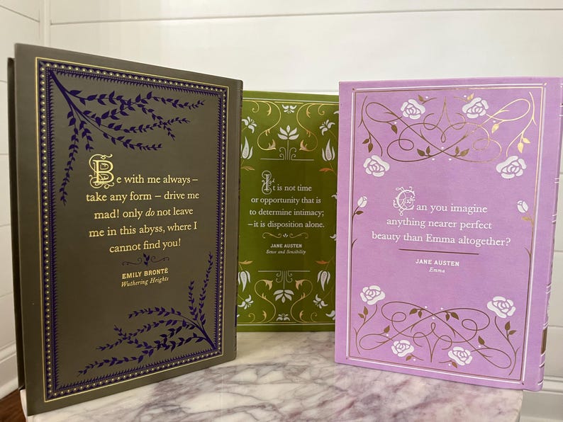 May include: Three hardcover books with gold lettering and decorative floral designs on the covers. The book on the left has a dark green cover with a quote from Emily Bront&euml;'s "Wuthering Heights." The middle book has a green cover with a quote from Jane Austen's "Sense and Sensibility." The book on the right has a light purple cover with a quote from Jane Austen's "Emma."