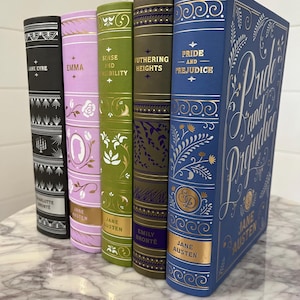 May include: A collection of six classic novels with gold lettering and decorative designs on the spines. The books are arranged from left to right: Jane Eyre, Emma, Sense and Sensibility, Wuthering Heights, Pride and Prejudice, and Pride and Prejudice.