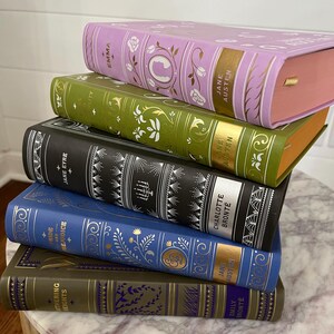 May include: A stack of six classic literature books with gold lettering and floral designs on the covers. The books are arranged from largest to smallest, with the top book being "Emma" by Jane Austen. The other books are "Pride and Prejudice" by Jane Austen, "Jane Eyre" by Charlotte Bronte, "Wuthering Heights" by Emily Bronte, "Sense and Sensibility" by Jane Austen, and "The Scarlet Letter" by Nathaniel Hawthorne.