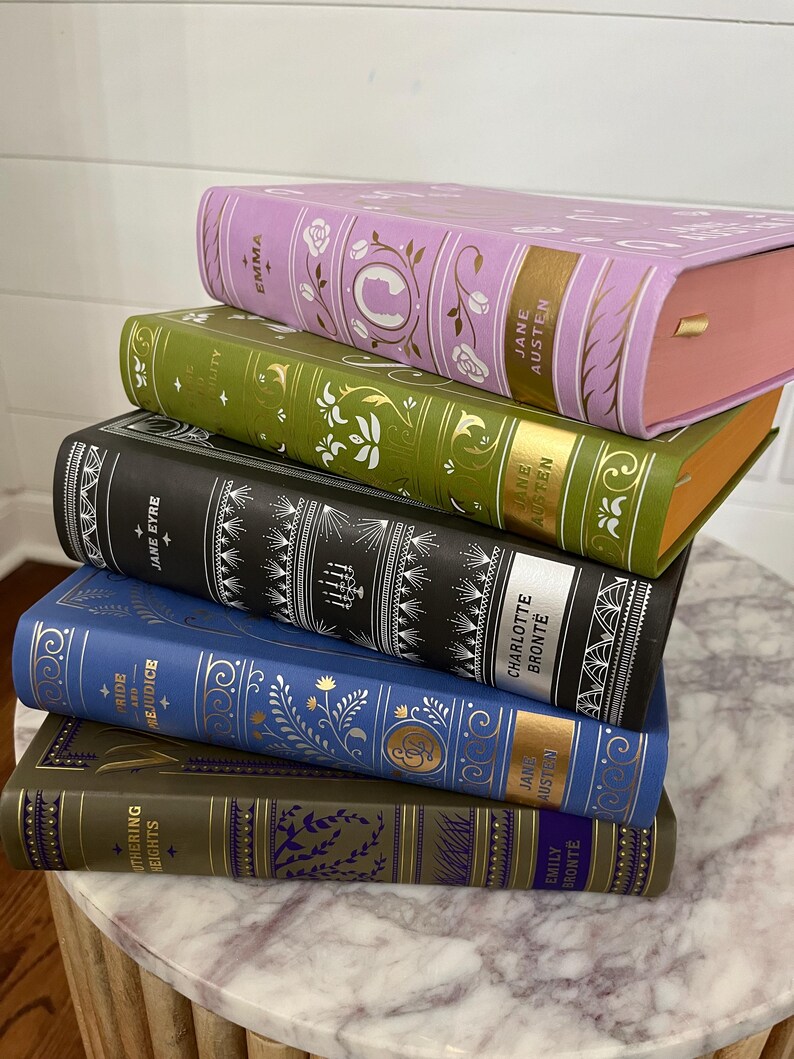 May include: A stack of six classic literature books with gold lettering and floral designs on the covers. The books are arranged from largest to smallest, with the top book being "Emma" by Jane Austen. The other books are "Pride and Prejudice" by Jane Austen, "Jane Eyre" by Charlotte Bronte, "Wuthering Heights" by Emily Bronte, "Sense and Sensibility" by Jane Austen, and "The Scarlet Letter" by Nathaniel Hawthorne.