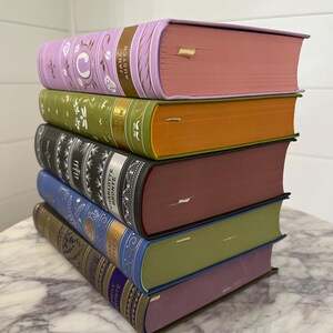 May include: A stack of six hardcover books with different colored covers. The top book is lavender with gold lettering that reads "Jane Austen Emma". The second book is green with gold lettering that reads "Pride and Prejudice". The third book is black with gold lettering that reads "Charlotte Bronte Jane Eyre". The fourth book is burgundy with gold lettering that reads "Villette". The fifth book is blue with gold lettering that reads "Emily Bronte Wuthering Heights". The bottom book is purple with gold lettering that reads "Jane Eyre".
