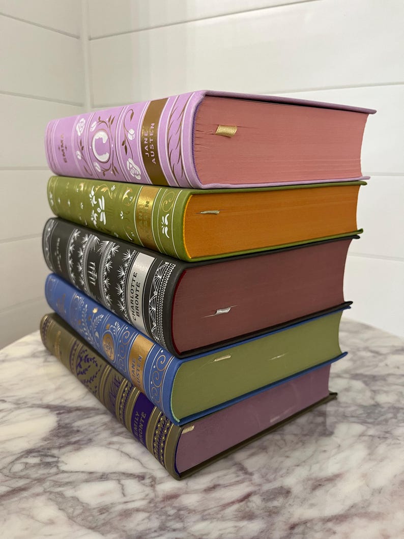 May include: A stack of six hardcover books with different colored covers. The top book is lavender with gold lettering that reads "Jane Austen Emma". The second book is green with gold lettering that reads "Pride and Prejudice". The third book is black with gold lettering that reads "Charlotte Bronte Jane Eyre". The fourth book is burgundy with gold lettering that reads "Villette". The fifth book is blue with gold lettering that reads "Emily Bronte Wuthering Heights". The bottom book is purple with gold lettering that reads "Jane Eyre".