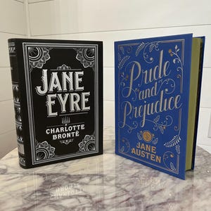 May include: Two hardcover books with decorative covers. The book on the left is black with silver lettering and says "Jane Eyre" by Charlotte Bronte. The book on the right is blue with silver lettering and says "Pride and Prejudice" by Jane Austen.