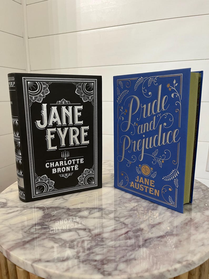 May include: Two hardcover books with decorative covers. The book on the left is black with silver lettering and says "Jane Eyre" by Charlotte Bronte. The book on the right is blue with silver lettering and says "Pride and Prejudice" by Jane Austen.