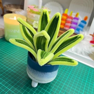 May include: A decorative plant made from green and yellow pipe cleaners, presented in a small, blue and white ceramic pot with three white feet. The plant has a vibrant, textured appearance, resembling a stylized succulent.