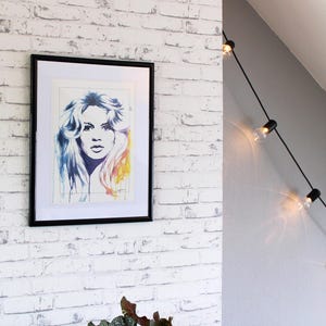 Peut inclure: Un portrait encadr&eacute; &agrave; l'aquarelle d'une femme aux longs cheveux flottants. L'&oelig;uvre pr&eacute;sente une palette de couleurs bleues, jaunes et oranges. Le cadre noir est mont&eacute; sur un mur de briques blanches. Des guirlandes lumineuses d&eacute;coratives sont visibles en arri&egrave;re-plan.