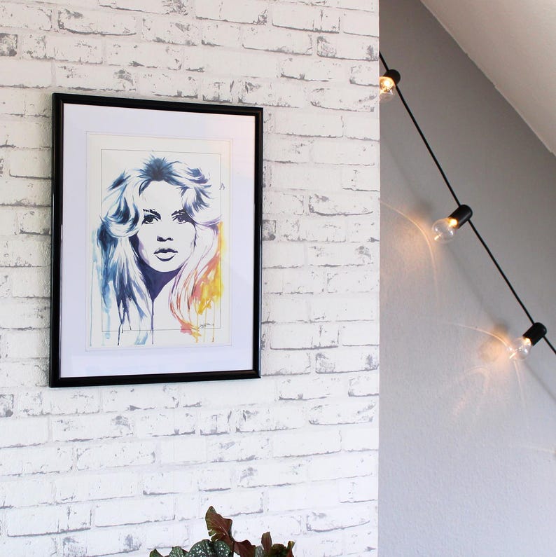 Peut inclure: Un portrait encadr&eacute; &agrave; l'aquarelle d'une femme aux longs cheveux flottants. L'&oelig;uvre pr&eacute;sente une palette de couleurs bleues, jaunes et oranges. Le cadre noir est mont&eacute; sur un mur de briques blanches. Des guirlandes lumineuses d&eacute;coratives sont visibles en arri&egrave;re-plan.