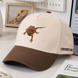 May include: A cream-colored baseball cap with a brown brim, featuring an embroidered bearded dragon on the front panel and the text "CHUNKY FAN" printed in black on the side. The cap has a small brown button on the top center of the crown.