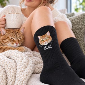May include: A pair of black crew socks featuring a graphic illustration of an orange tabby cat face and the name ARCHIE printed in white capital letters below the cat's chin. The socks are made of a ribbed knit fabric and reach mid-calf.