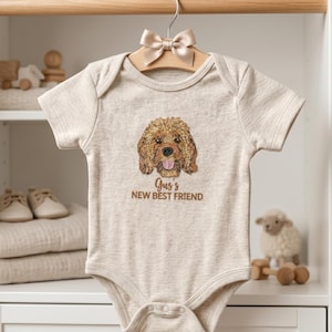 May include: A beige short-sleeved baby bodysuit featuring an embroidered golden-brown curly-haired dog face with a pink tongue. Below the dog, the text reads Gus's NEW BEST FRIEND in brown embroidery. The garment hangs from a wooden hanger.