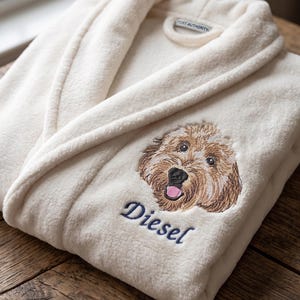 Könnte beinhalten: Cremefarbener Plüschbademantel mit einem gestickten Hundegesicht und dem Namen "Diesel" in marineblauer Schrift. Der Bademantel hat einen Schalkragen und eine Schlaufe zum Aufhängen. Das Etikett lautet "Port Authority."