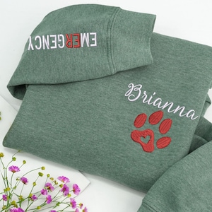 Peut inclure: Un sweat-shirt vert avec le mot "EMERGENCY" brodé en blanc et rouge sur la manche. Le devant présente le nom "Brianna" en écriture blanche et une empreinte de patte rouge avec un cœur.
