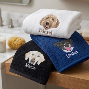 May include: A stack of three folded terry cloth towels in white, navy blue, and black. Each towel features an embroidered dog portrait and a name: a golden-brown dog named Diesel, a black and white dog named Cooper, and a cream dog named Macie.