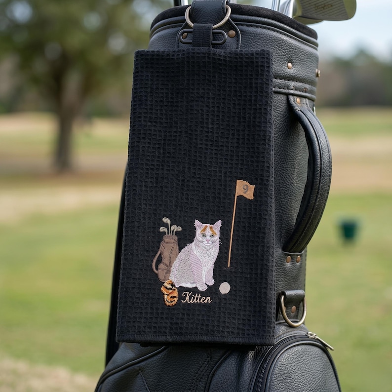 K&ouml;nnte beinhalten: Schwarzes Golfhandtuch mit einem gestickten Design mit einer Katze, Golfschl&auml;gern, einem Golfball und einer Fahne mit der Nummer 9. Das Wort "Kitten" ist unter der Katze gestickt. Das Handtuch h&auml;ngt an einer schwarzen Golftasche.