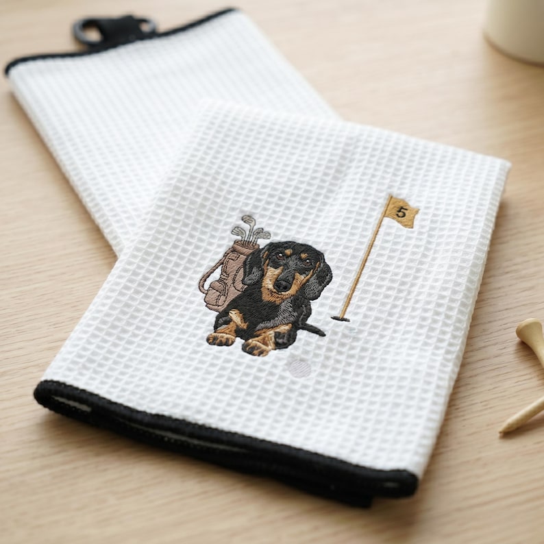 K&ouml;nnte beinhalten: Wei&szlig;es Golfhandtuch mit schwarzem Rand und Waffelstruktur. Das Handtuch zeigt einen gestickten Dackel mit Golftasche und einer Flagge mit der Nummer 5. Das Handtuch hat einen schwarzen Clip zum Aufh&auml;ngen.