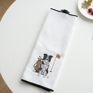 K&ouml;nnte beinhalten: Wei&szlig;es Golfhandtuch mit schwarzem Rand und Waffelstruktur. Das Handtuch hat ein gesticktes Design eines schwarz-wei&szlig;en Hundes, einer Golftasche und einer Flagge mit der Nummer 9.