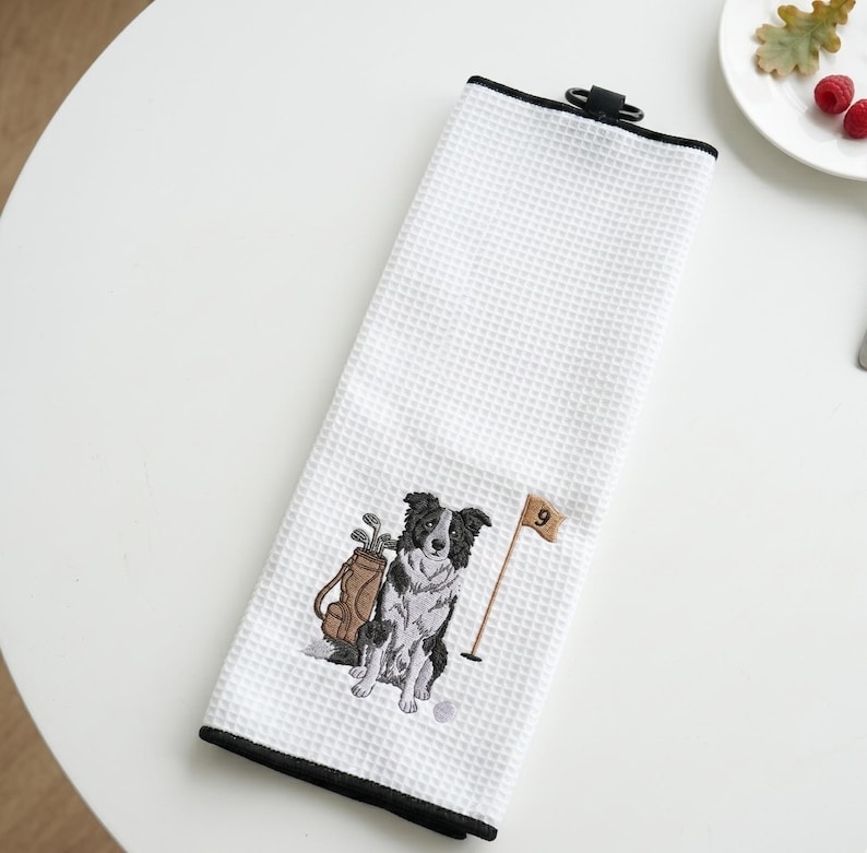 K&ouml;nnte beinhalten: Wei&szlig;es Golfhandtuch mit schwarzem Rand und Waffelstruktur. Das Handtuch hat ein gesticktes Design eines schwarz-wei&szlig;en Hundes, einer Golftasche und einer Flagge mit der Nummer 9.