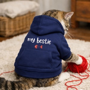 Può includere: Un gatto tigrato indossa una felpa con cappuccio blu navy con la scritta "my bestie" e un disegno a lisca di pesce e cuore. Il gatto sta giocando con una palla di filo rossa. La felpa ha una tasca.
