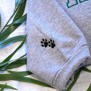 Sport Grey embroidered dog lover sweatshirt sleeve featuring two small paw print stitches as a minimalist pet detail. The soft cotton sweatshirt fabric and clean embroidery make this custom pet portrait sweatshirt stylish and sentimental. This personalized dog sweatshirt pairs perfectly with custom dog face embroidery on the chest, creating a thoughtful gift for dog moms, dog dads, and pet lovers.
