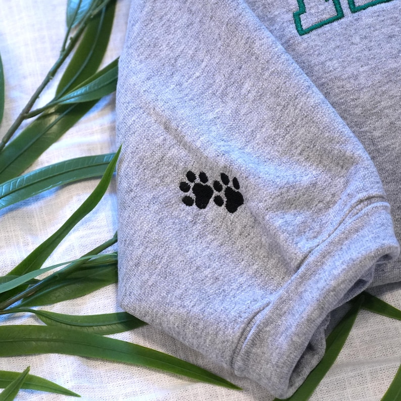 Sport Grey embroidered dog lover sweatshirt sleeve featuring two small paw print stitches as a minimalist pet detail. The soft cotton sweatshirt fabric and clean embroidery make this custom pet portrait sweatshirt stylish and sentimental. This personalized dog sweatshirt pairs perfectly with custom dog face embroidery on the chest, creating a thoughtful gift for dog moms, dog dads, and pet lovers.