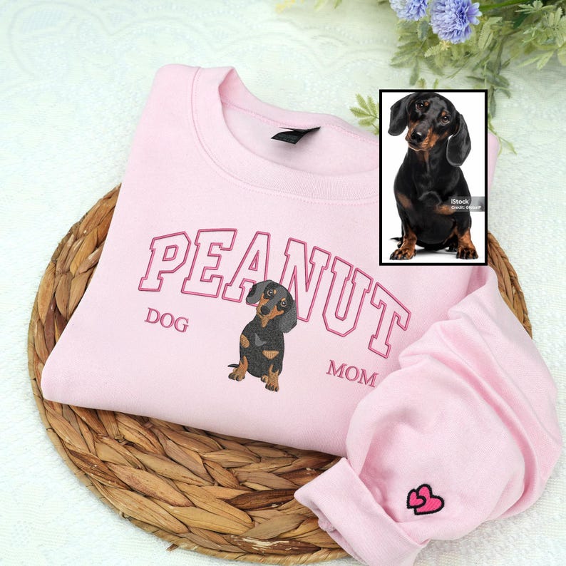 Light pink embroidered sweatshirt with “PEANUT DOG MOM” text and a lifelike black-and-tan dachshund portrait. Detailed embroidery with pink varsity letters and a small heart on the sleeve — personalized embroidered gift for dog moms.