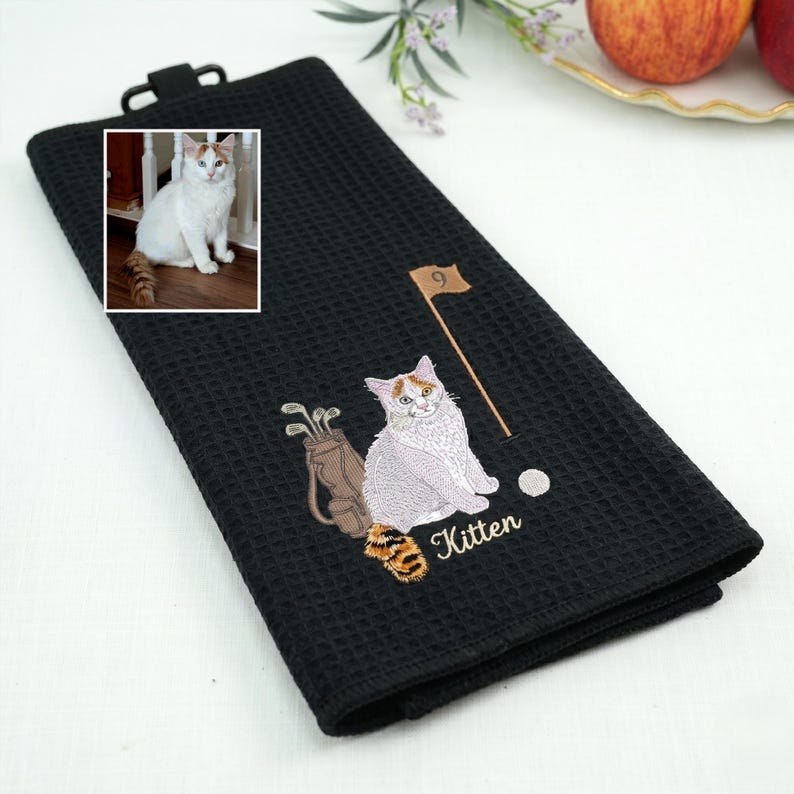 K&ouml;nnte beinhalten: Schwarzes Golfhandtuch mit Waffelstruktur. Es zeigt ein gesticktes Design einer wei&szlig;-orangenen Katze, Golfschl&auml;gern, einem Golfball und einer Fahne. Das Wort "Kitten" ist darunter gestickt.