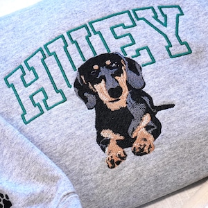 Close-up of a custom dachshund embroidery on a Sport grey sweatshirt with the name “Huey” stitched in varsity style lettering above the dog portrait. The detailed embroidery highlights the texture of the stitches and realistic pet illustration inspired by the customer’s dog photo. This custom dog portrait sweatshirt is a meaningful personalized gift for dog lovers, dachshund owners, and pet parents.