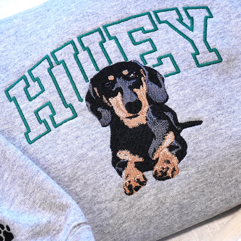 Close-up of a custom dachshund embroidery on a Sport grey sweatshirt with the name “Huey” stitched in varsity style lettering above the dog portrait. The detailed embroidery highlights the texture of the stitches and realistic pet illustration inspired by the customer’s dog photo. This custom dog portrait sweatshirt is a meaningful personalized gift for dog lovers, dachshund owners, and pet parents.