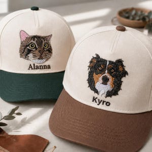 Puede incluir: Dos gorras de béisbol con coronas color crema. La gorra izquierda tiene una visera verde oscuro y un gato atigrado bordado con el nombre Alanna. La gorra derecha tiene una visera marrón y un perro tricolor bordado con el nombre Kyro.