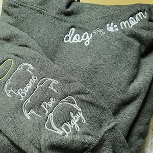May include: Dark gray hoodie with white embroidered text and dog illustrations. The hoodie features the words "dog mom" and the names "Boone", "Poe", and "Digby" embroidered on the sleeve.