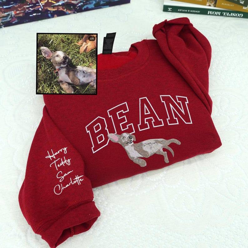 Red embroidered sweatshirt featuring a tan-and-white dog portrait and large varsity text “BEAN” across the chest. Left sleeve personalized with embroidered names Harry, Teddy, Sam, Charlotte in white thread — heartfelt custom pet lover sweatshirt.