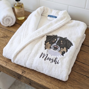 May include: White terry cloth bathrobe with an embroidered dog face and the name "Mooshi" in script. The robe has a shawl collar and is folded on a wooden surface. A rolled-up white towel and a bottle of bath oil are in the background.