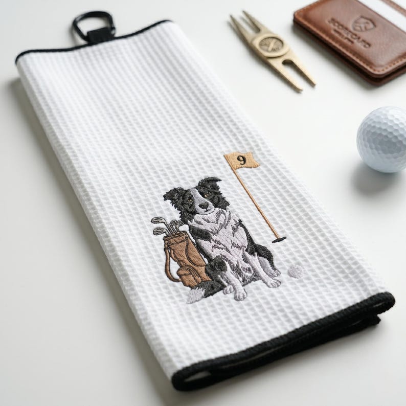K&ouml;nnte beinhalten: Wei&szlig;es Golfhandtuch mit schwarzem Rand und Aufh&auml;ngeschlaufe. Das Handtuch zeigt ein gesticktes Bild eines Border Collie-Hundes, einer Golftasche und einer Flagge mit der Nummer 9. Ein Golfball, ein Pitchgabel und ein Lederkartenhalter sind ebenfalls zu sehen.
