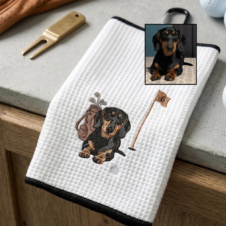 K&ouml;nnte beinhalten: Wei&szlig;es Golfhandtuch mit schwarzem Rand und einer schwarz-braunen Dackel-Stickerei. Die Stickerei umfasst eine Golftasche, eine Golf-Flagge und ein Dackel-Portr&auml;t. Ein Golf-Tee und ein Golfhandschuh befinden sich im Hintergrund.
