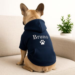 Könnte beinhalten: Ein marineblauer Hunde-Hoodie mit dem Namen "Bruno" in weißer Schrift und einem weißen Pfotenabdruck. Der Hoodie hat eine Kapuze und eine Vordertasche. Der Hund ist eine hellbraune französische Bulldogge.