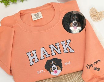 May include: A peach-colored crewneck sweatshirt featuring embroidered text that reads HANK, with the year 2015 and the text EST. surrounding an embroidered dog face. The sleeve features the embroidered text Dog mom with paw prints.