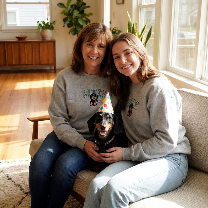 Mother and daughter wearing matching Sport grey custom dog portrait sweatshirts while sitting on a sofa with their dachshund wearing a birthday hat. The embroidered sweatshirt features the name “Huey” and a dachshund portrait design. This personalized dog lover sweatshirt is a heartwarming gift idea for pet parents, dog moms, family pet lovers, and custom pet portrait apparel.