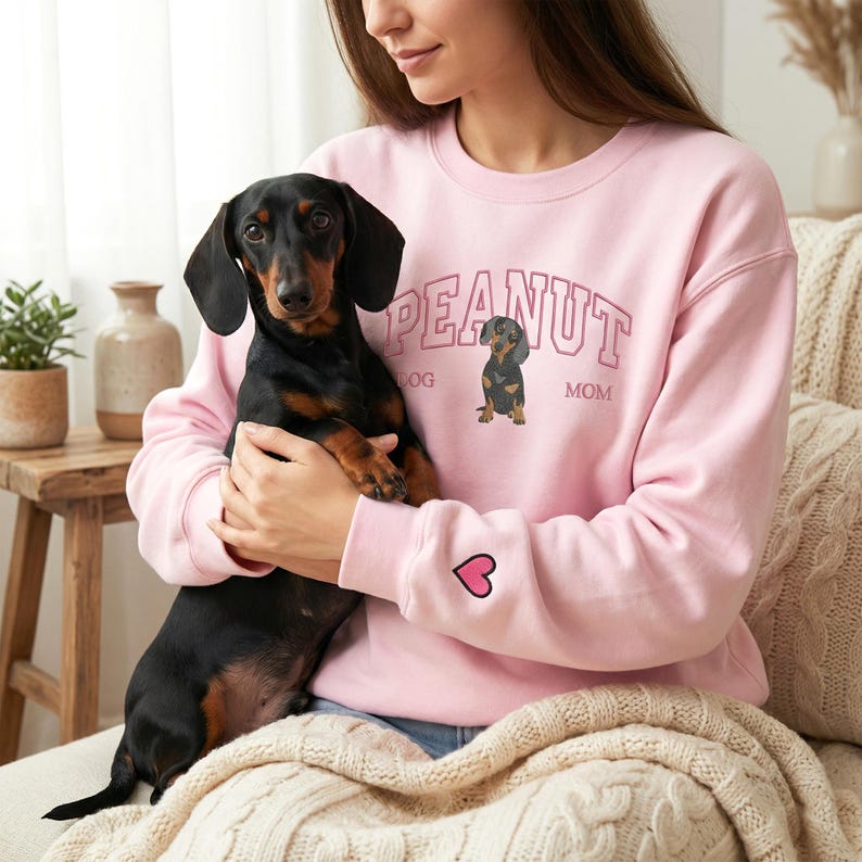 Woman holding a black and tan dachshund while wearing a light pink sweatshirt with custom dog illustration and text “PEANUT Dog Mom” embroidered on the chest and a small heart detail on the sleeve. Warm indoor lifestyle setting with soft blankets and natural light. Personalized dog mom sweatshirt, dachshund gift, custom pet apparel, cozy handmade dog lover gift.